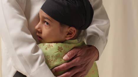 close up studio shot of young sikh boy with top knot turban hugging grandfather against plain background 2