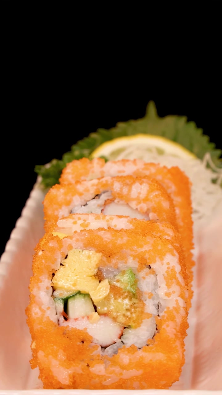 Yksityiskohtainen lähikuva California roll -sushista pyörii valkoisella lautasella mustaa taustaa vasten, kirkkaan valaistuksen korostaessa eloisia värejä ja rakenteita.