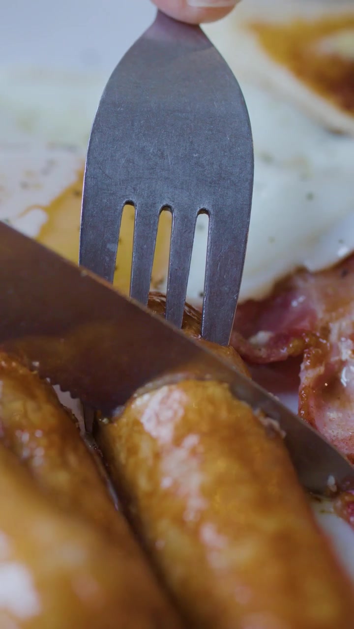 Primo piano di forchetta e coltello che tagliano la salsiccia accanto a pancetta e uovo, luce naturale, messa a fuoco ridotta
