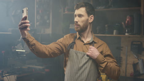 young caucasian male carpenter in apron holding smartphone and making a selfie with mobile phone