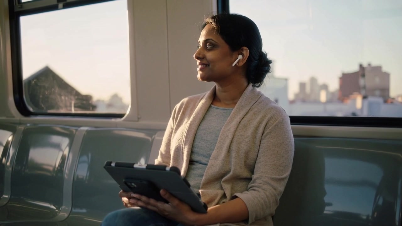 Noticing passing buildings, woman in beige cardigan turning to window on train, tablet and earbuds