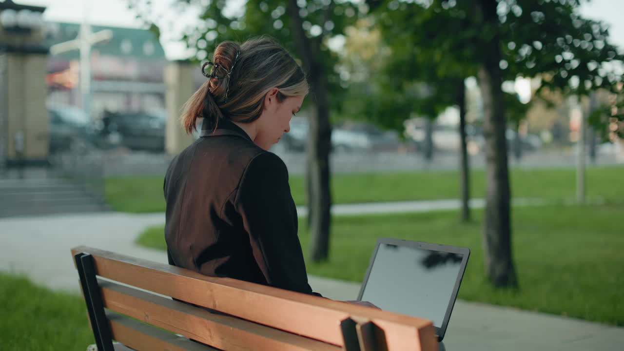 Side view of lady typing on laptop seated on wooden bench in park surrounded by lush greenery with distant parked cars and urban structures visible in blurred background