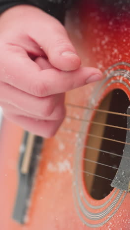 primer plano de las cuerdas de la guitarra acústica tocadas a mano en polvo de nieve, mostrando vibrantes tonos de guitarra rojo-naranja y detalles esmaltados, una armonía perfecta entre la música y el invierno