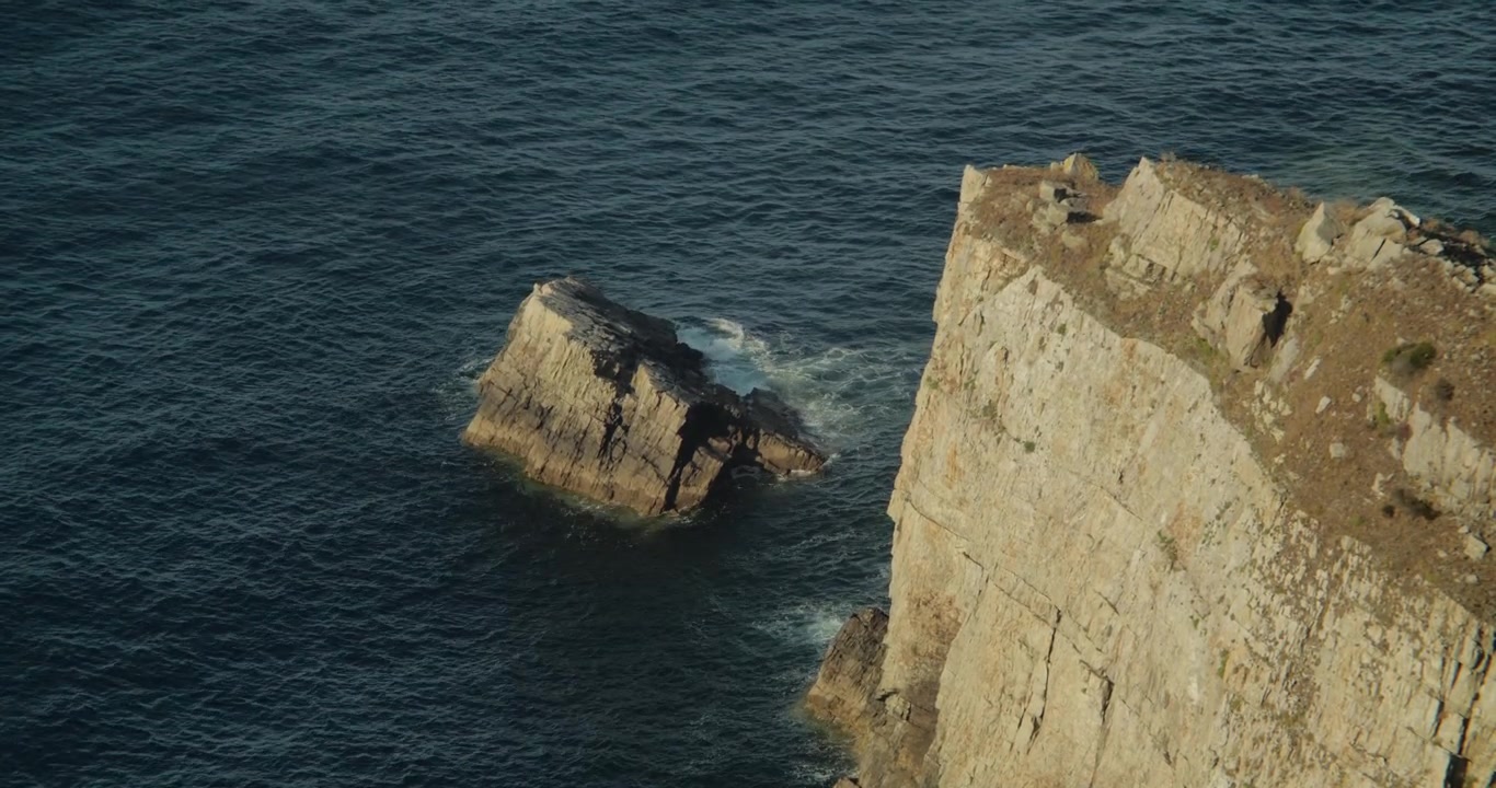 Handheld horizontal shot captures rugged Cabo Peñas cliffs, textured rocks, native coastal plants, and calm Atlantic Ocean waves in golden natural light, evoking serene wild beauty.