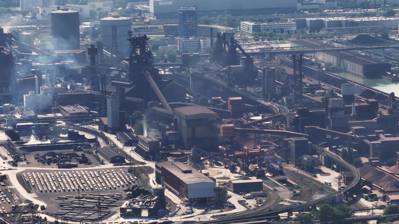 High angle view of a large steel plant with blast furnaces, chimneys, and industrial buildings. A freight train maneuvers within the complex