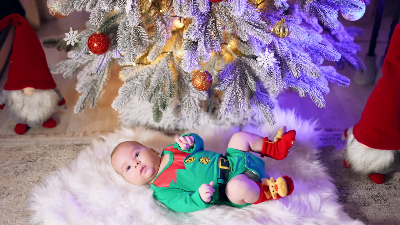 Caucasian child in green elf costume and red socks with deer resting on the fluffy plaid. Beautiful toddler moving legs actively under Christmas tree.