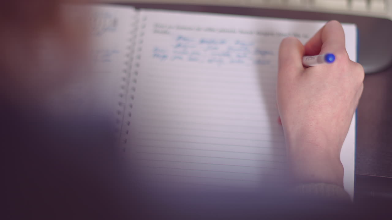 Close up of a woman's hand writing in a spiral notebook with a blue pen while sitting at a desk