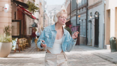 mujer rubia muy contenta con auriculares inalámbricos eligiendo escuchar música sosteniendo un teléfono inteligente en la calle