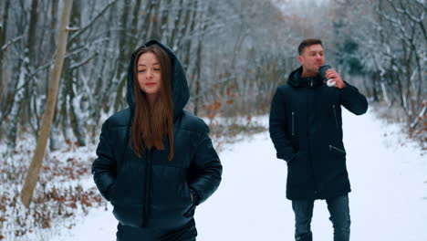 man and woman walking in winter nature. woman walks ahead holding her hands in pockets. man walks behind drinking coffee.