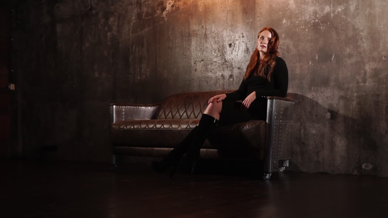 una mujer con un vestido negro sentada en un sofá de cuero en un entorno industrial oscuro.