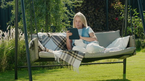 una mujer linda usa una tableta. descansando en un columpio de jardín en el patio trasero de una casa