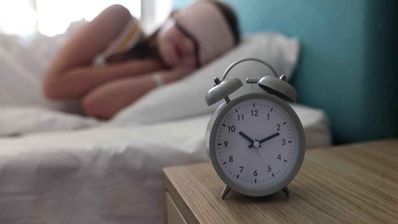 Alarm clock on a nightstand with a person sleeping in the background