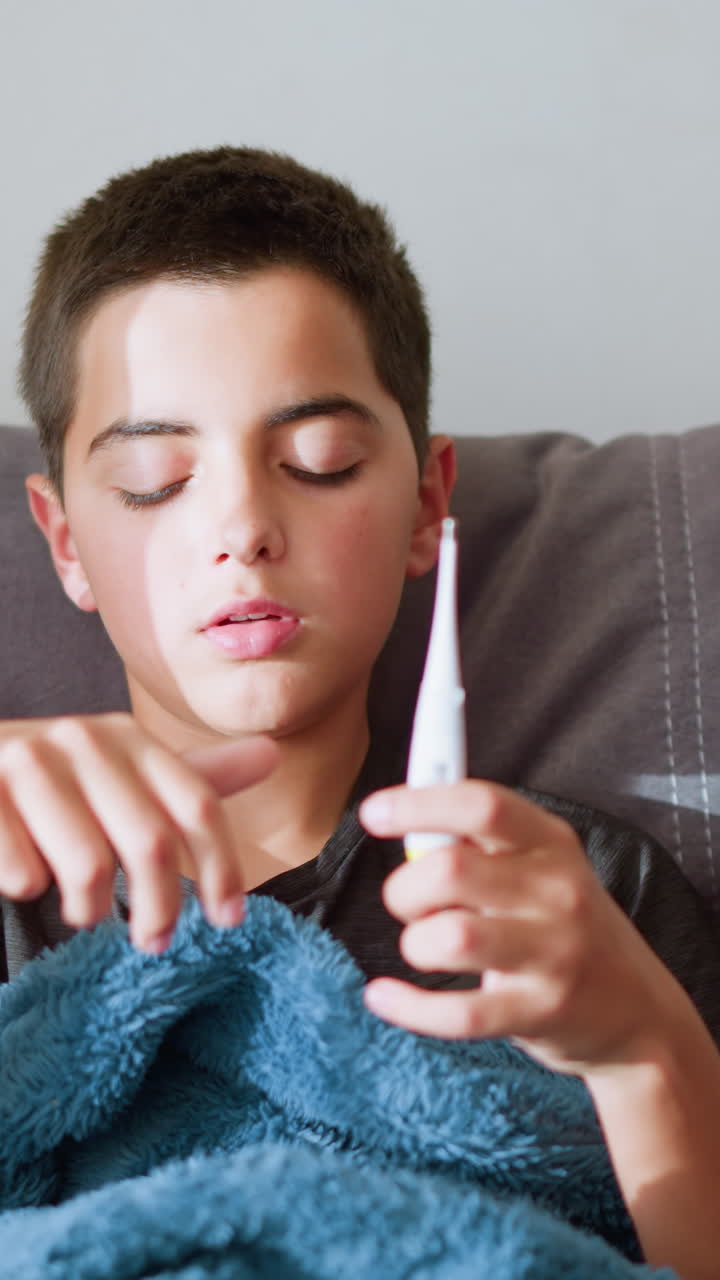 Ragazzo malato seduto sul divano che allunga la mano per ricevere il termometro dal fratello/sorella per controllare la temperatura mentre è coperto da una coperta calda, la luce naturale filtra attraverso la finestra