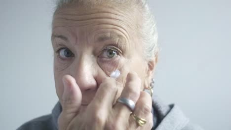 Blue-eyed-caucasian-senior-woman-looks-at-camera-putting-cream-at-face-closeup-portrait-shot