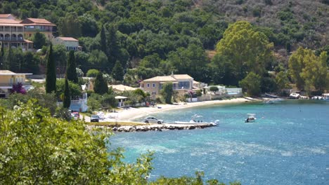 Vista-De-Una-Playa-Junto-Al-Mar-Azul-En-La-Isla-De-Corfú,-Con-Edificios-Y-Apartamentos-Al-Fondo