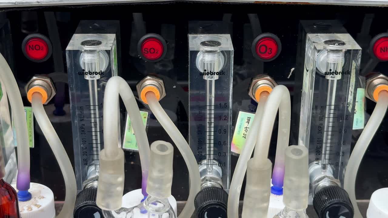 Laboratory gas analysis system with labeled flow meters for NO₂, SO₂, and O₃, transparent tubing, and chemical absorption bottles used for environmental air quality testing. Impinger machine.