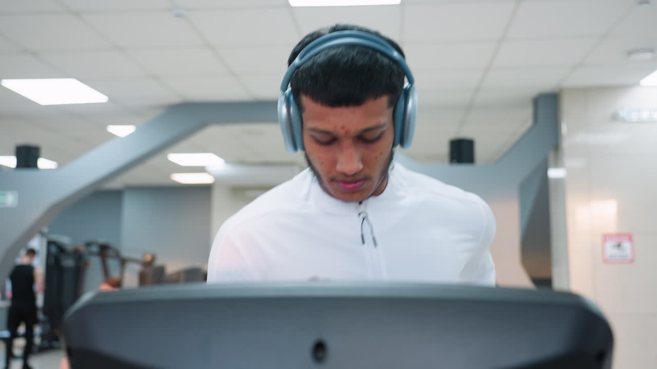 close up of determined man in white top running on treadmill in modern gym under bright lights, focused expression, blurred background shows equipment, windows and urban view beyond glass