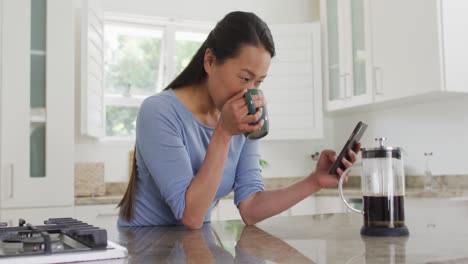 Feliz-Mujer-Asiática-Parada-En-La-Mesa,-Tomando-Café-Y-Usando-Un-Teléfono-Inteligente-En-La-Cocina