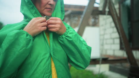 close-up of a woman buttoning her green raincoat outdoors next to swing bench, with blurred background featuring greenery, two building and window