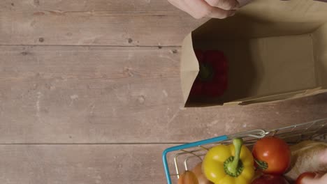 overhead shot of person packing basic fresh food items from supermarket wire shopping basket into paper bag