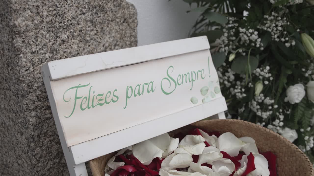 White wooden wedding sign with script text beside rose petals and gypsophila at stone wall