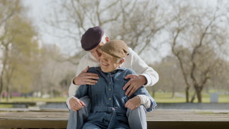 lovely senior couple looking at each other and kissing while sitting on bench in park at pond on nice autumn day