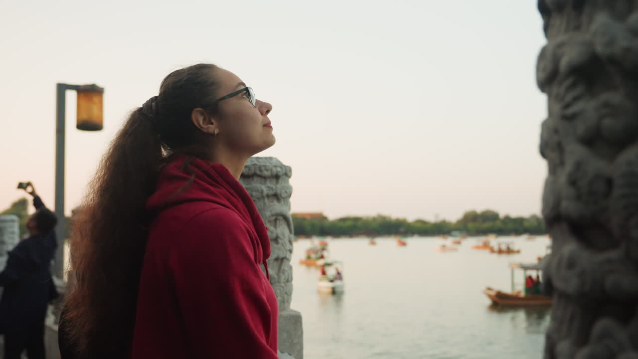 Giovane donna con felpa rossa al tramonto in riva al lago, in piedi accanto a un pilastro di pietra intagliata presso un padiglione storico, con lo zaino in spalla, occhiali, lunghi capelli ricci, barche che galleggiano sull'acqua calma, morbido crepuscolo dorato.