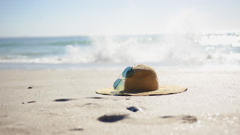 un sombrero de paja y gafas de sol descansan en una playa de arena, con el océano en el fondo, con espacio de copia