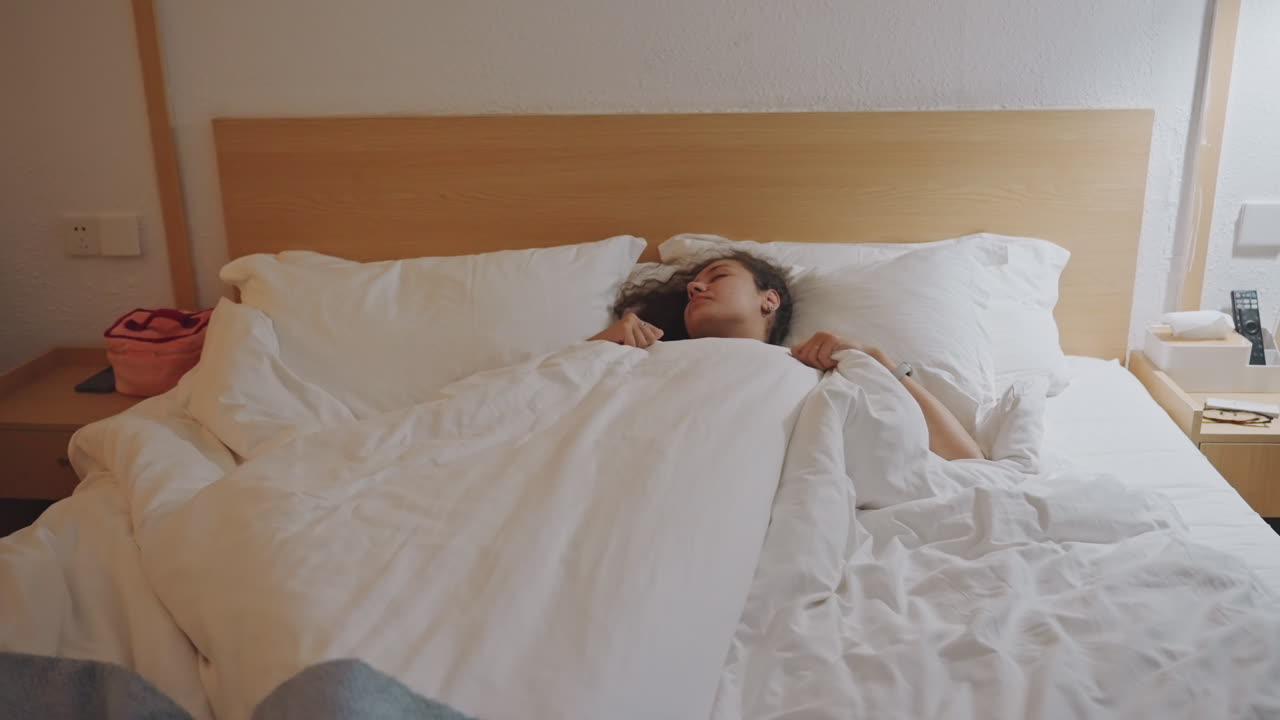 Asiatisk kvinde sover i hotelseng, blødt hvidt sengetøj og varmt lampelys. Første billede viser udmattet forretningsrejsende der tjekker telefonen, andet billede viser solo turist der strækker sig under dynen, senere.