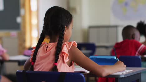 Portrait-of-happy-mixed-race-schoolgirl-sitting-at-classroom,-making-notes,-looking-at-camera