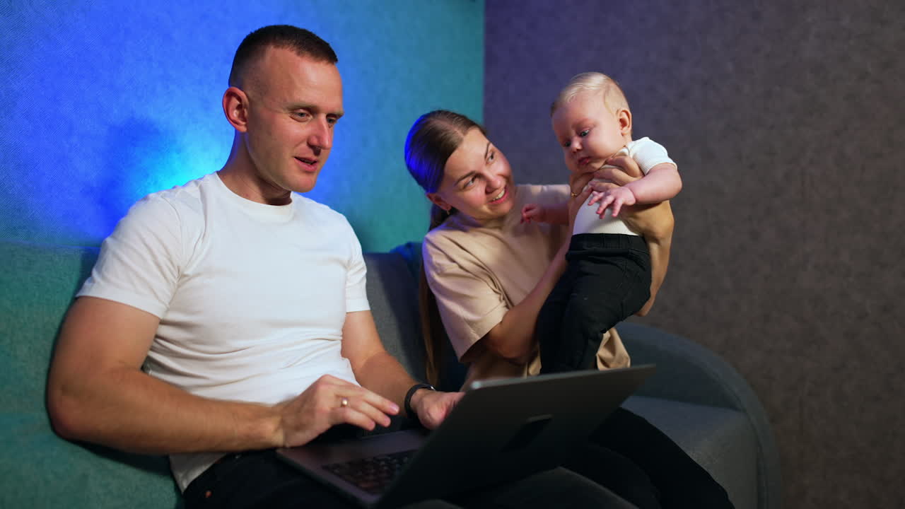 Caucasian man works on computer laptop. Wife sits beside holding a lovely blond infant baby.