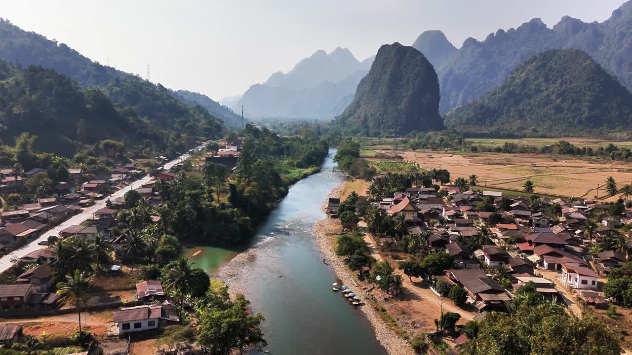 Aerial view of Pha Tang in Vang Vieng, Laos, featuring a winding river, village houses, green trees, and limestone mountains under a clear sky creating a tranquil rural atmosphere