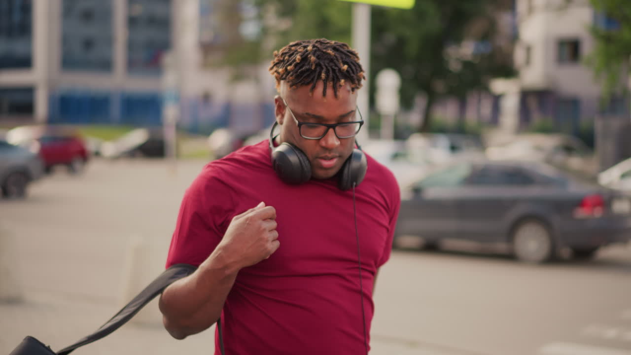 black man with headphones backpack adjusts gear on busy city sidewalk, scanning street, lifting case, readying instruments, wearing red tee and braided hair, urban cars and buildings in background