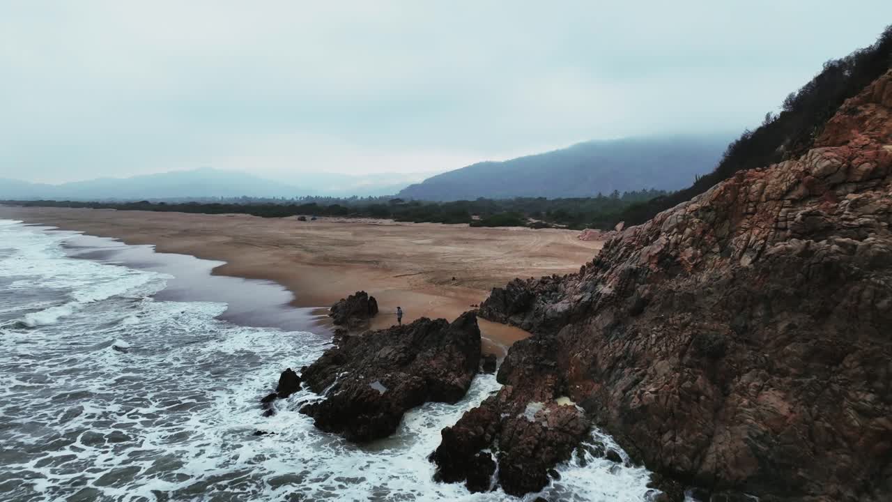Flyover powerful, foamy ocean waves at La Peña Blanca beach. Rocky cliffs on the shoreline and extensive golden sand on a cloudy day