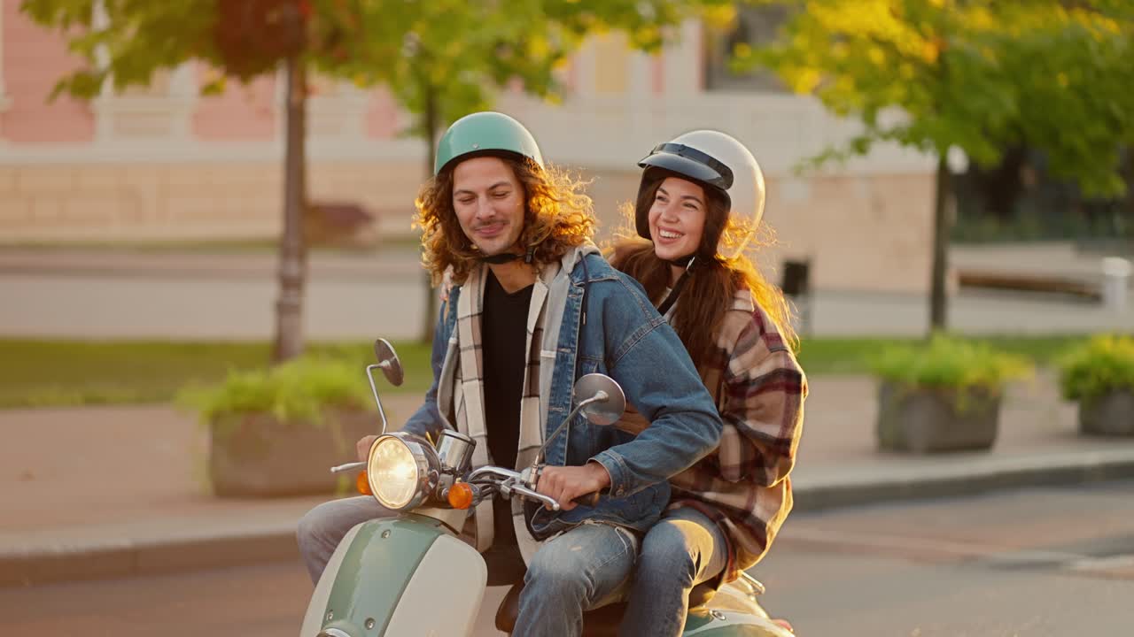 A happy couple, a guy with long curly hair in a denim jacket and a green moped helmet, rides with his brunette girlfriend in a checkered shirt and a white helmet on a green moped along a green summer city street