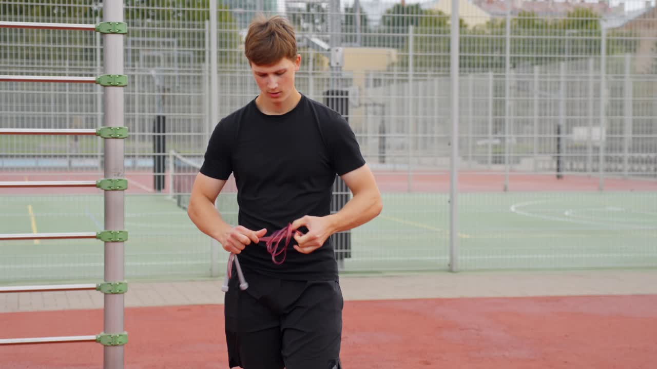 Athletic man ties jump rope and walks away post-workout, dressed in black sportswear, casual