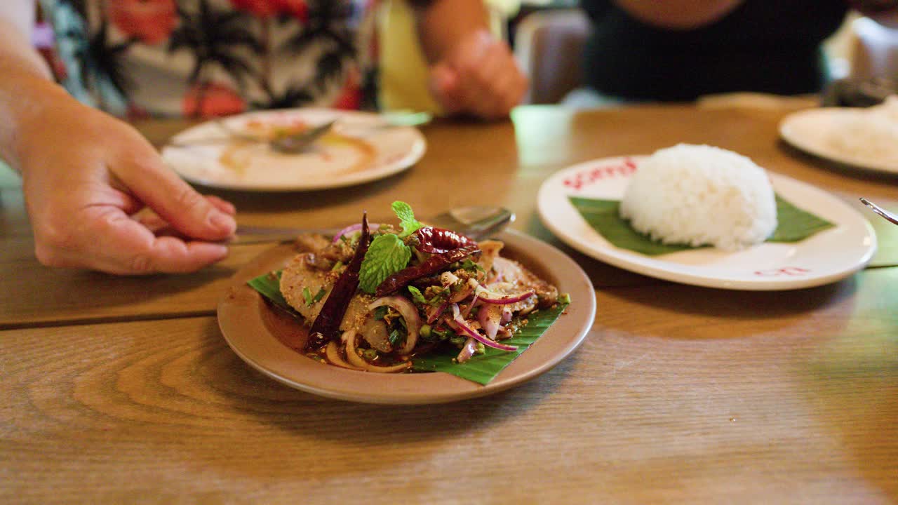 Hand serves spicy pork salad with herbs and rice in warmly lit Bangkok restaurant