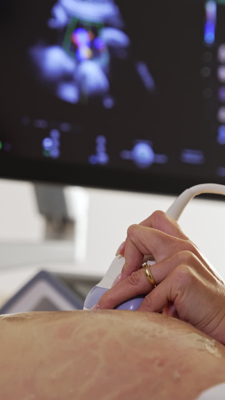Female doctor's hand moving the sensor by the big pregnant belly. Black screen of a sonographer at backdrop in blur. Vertical video