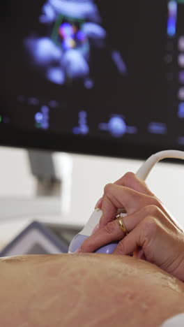 female doctor's hand moving the sensor by the big pregnant belly. black screen of a sonographer at backdrop in blur. vertical video