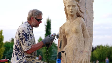 mature man creating a statue of woman from a wood. artist uses a grinding tool in his work.