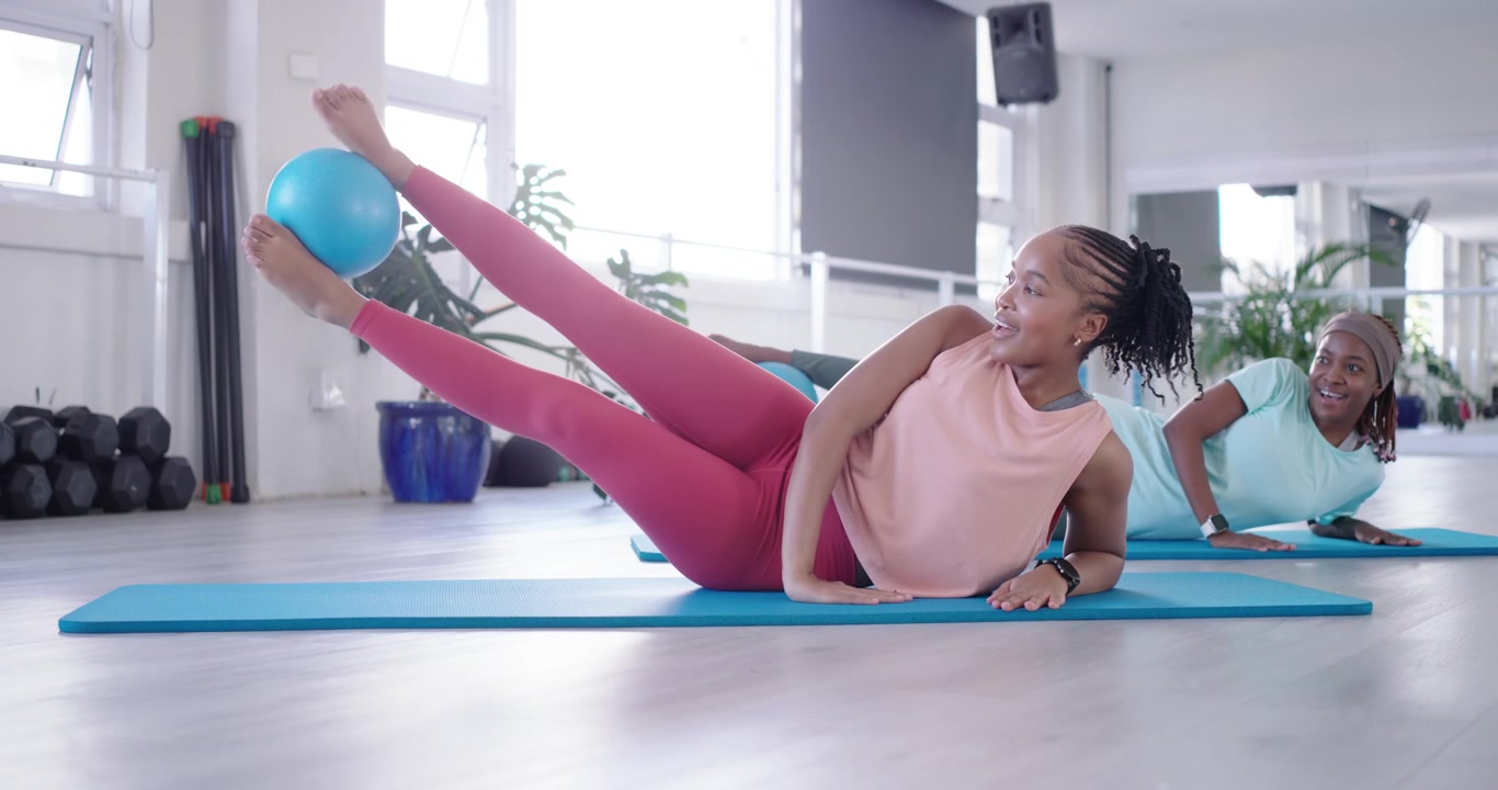 African American women in sportswear propping arms in gym, lifting legs, holding blue ball for core