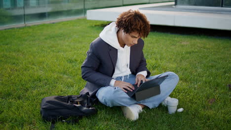 estudiante serio escribiendo portátil en el parque urbano. joven relajado sentado en la hierba