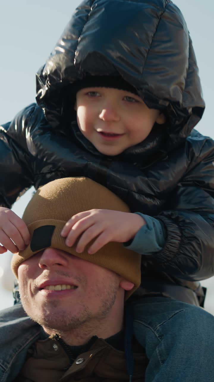 un primer plano de un padre llevando a su hijo pequeño en sus hombros, mientras el niño juguetón cubre la cara de su padre con un gorro, con una vista borrosa de los árboles detrás de ellos