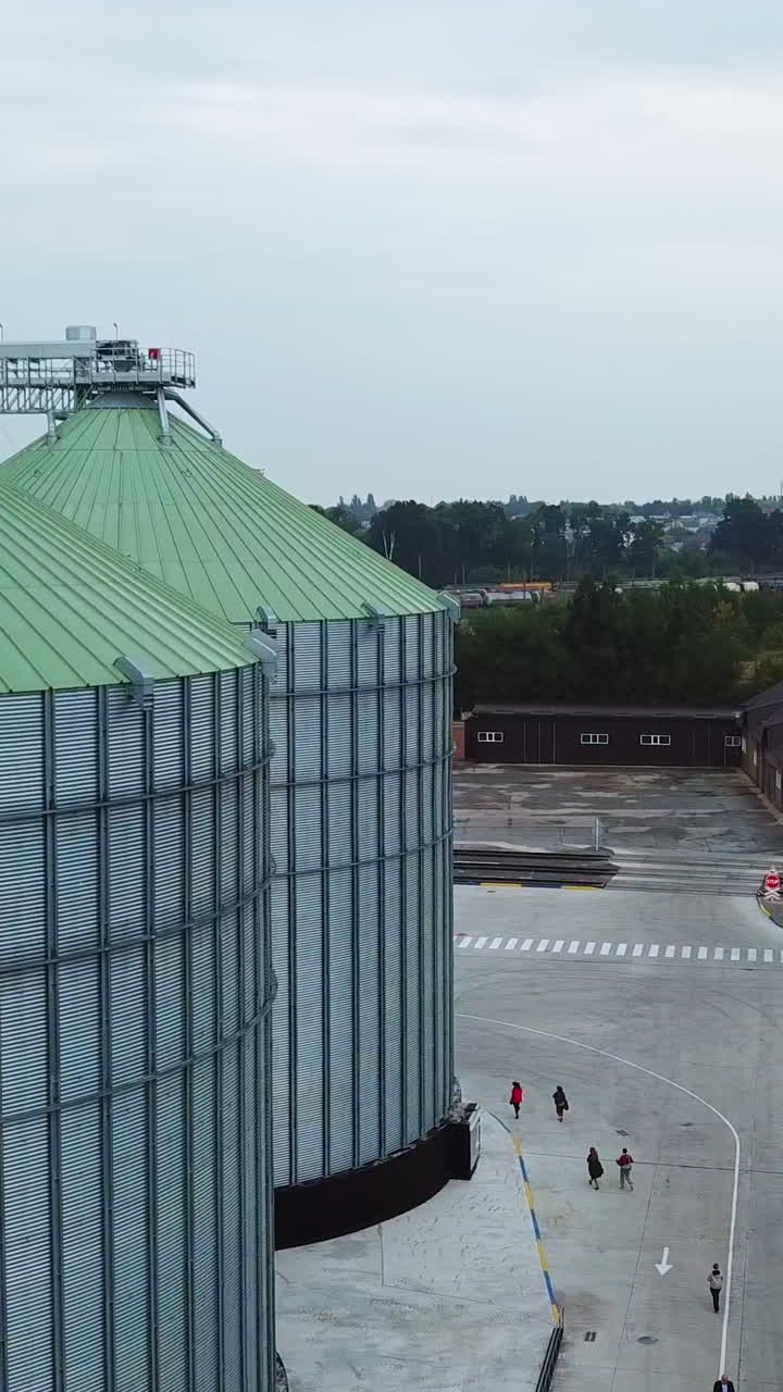 Metal tanks of elevator with diverse agricultural machinery nearby. People walking along the huge silver cisterns. View from above. Vertical video