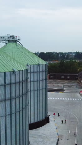 metal tanks of elevator with diverse agricultural machinery nearby. people walking along the huge silver cisterns. view from above. vertical video