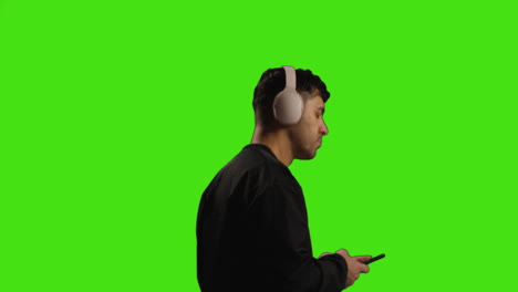 Young-Man-Wearing-Wireless-Headphones-And-Streaming-Music-Against-Green-Screen-With-Low-Key-Lighting-Walking-Across-Frame-1