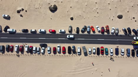 Vista-Estática-De-Arriba-Hacia-Abajo-De-Los-Autos-Estacionados-Junto-A-La-Carretera-Con-Autos-Lentos-Y-Turistas-Descansando-En-La-Arena-Amarilla-En-Un-Día-Caluroso