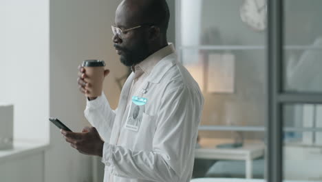 médico afroamericano bebiendo café y usando el teléfono en el trabajo