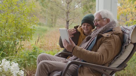 senior grandfather sitting with his grandson sitting on a bench in home garden while talking on a video call using a tablet