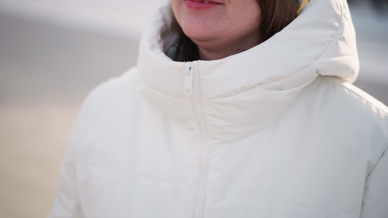 Cheerful young woman closing up zip on puffy winter coat with gloved hand while smiling warmly, black headphones resting around neck, winter light illuminating quilted fabric and rosy cheeks on day